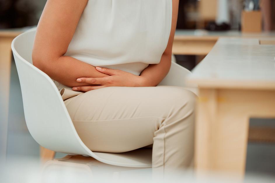 A photo of the lower body of a woman sitting in a chair clutching her stomach in pain. 