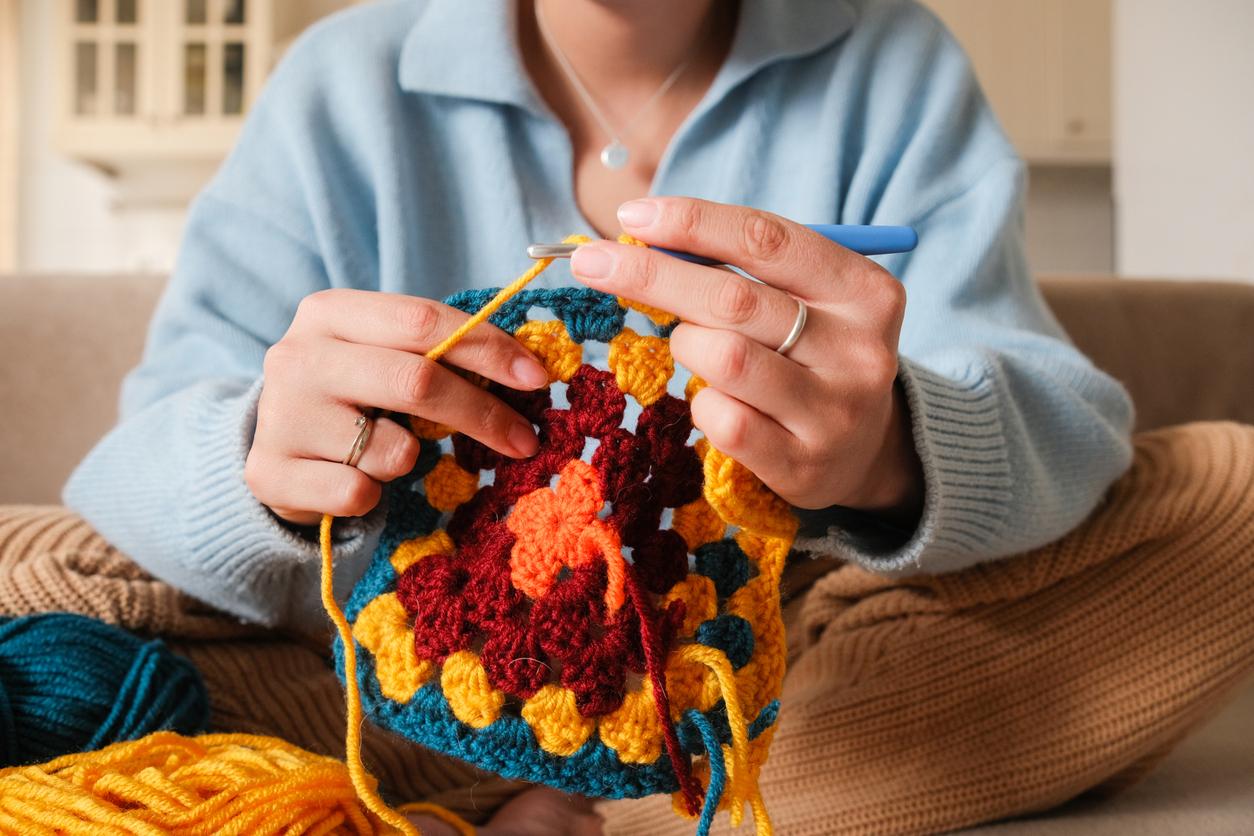 Close up of a person wearing a blue sweater crocheting a blue, yellow, red, and orange granny square.