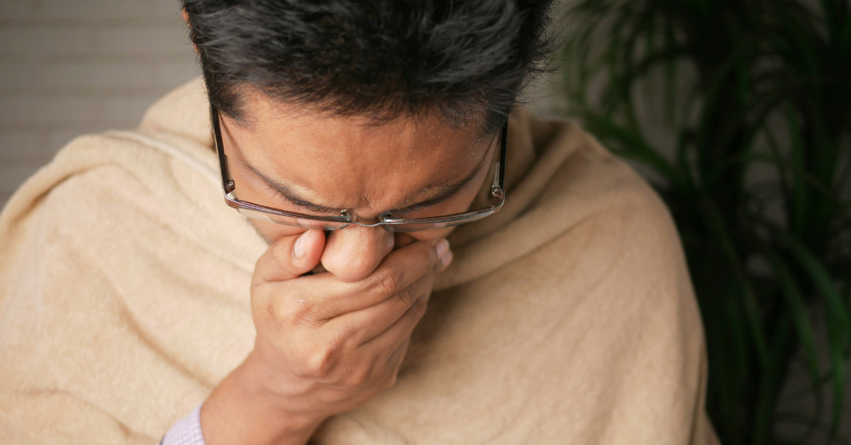 A man wrapped in a blanket coughs into his hand