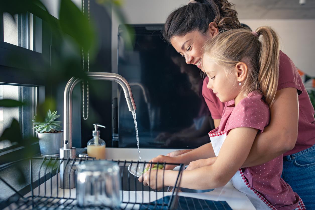 A smiling mother washes dishes with her daughter in the sink of their kitchen.