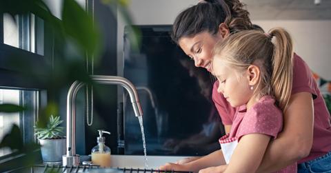 A smiling mother washes dishes with her daughter in the sink of their kitchen.