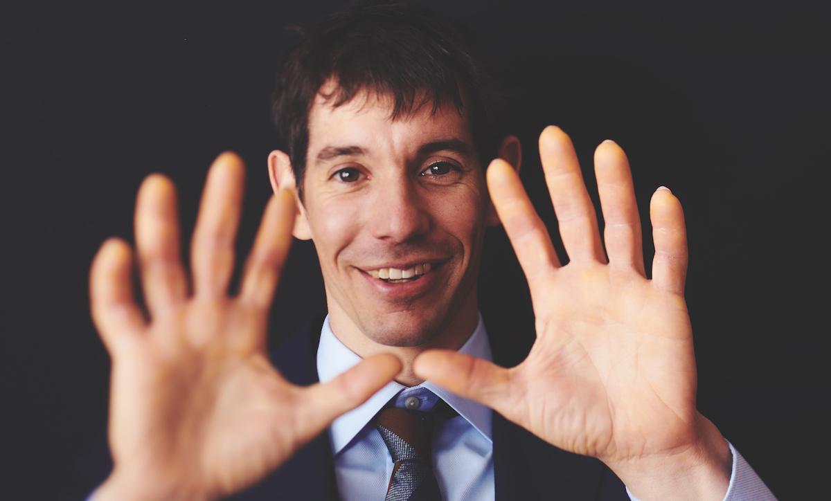 In front of a black background, Alex Honnold smiles at the camera, holding up his hands.