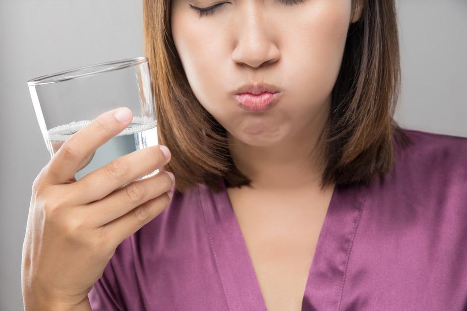 A woman wearing a purple shirt swishes water in her mouth while holding a glass. 