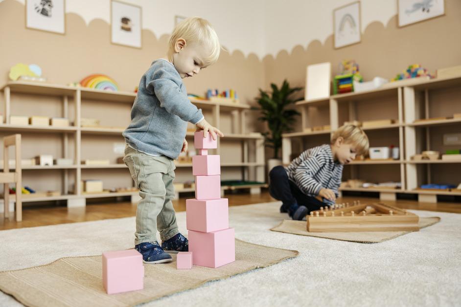 A child in the foreground stacks pink blocks in a tower while another child in the background plays with a wooden structure on the floor in a playroom of a preschool. 