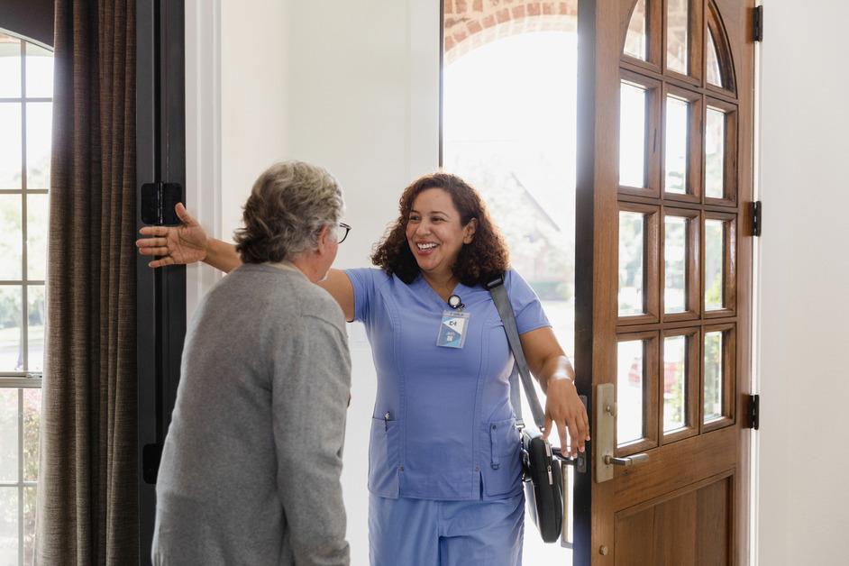 A nurse wearing blue scrubs walks through the door and holds out her arms to welcome a senior woman wearing a gray sweater