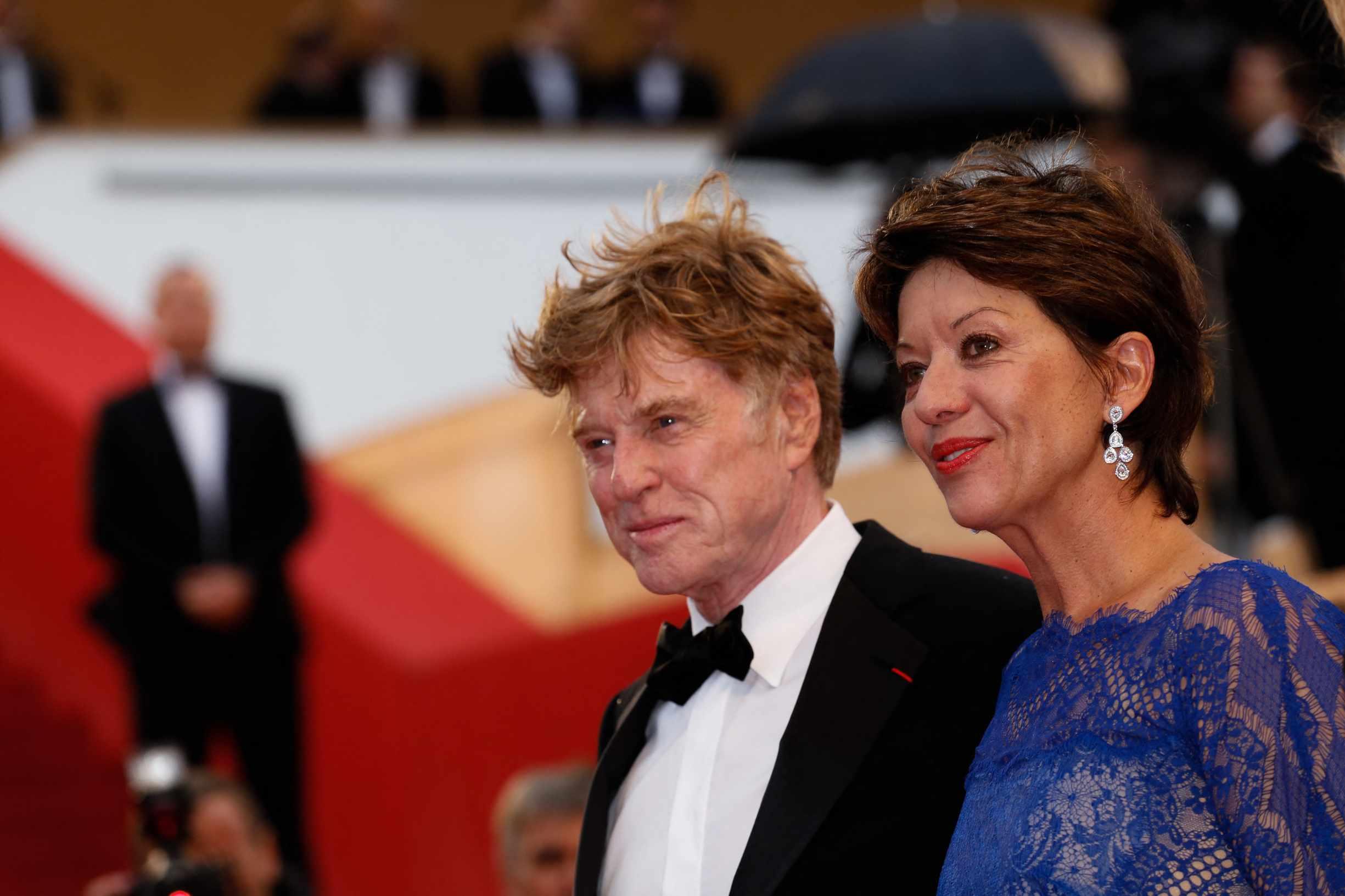 Robert Redford is pictured in a tuxedo while standing on the red carpet with a woman in a blue dress.