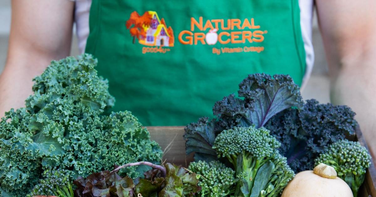 Man in Natural Grocers apron holds a wooden box of veggies, including kale and broccoli