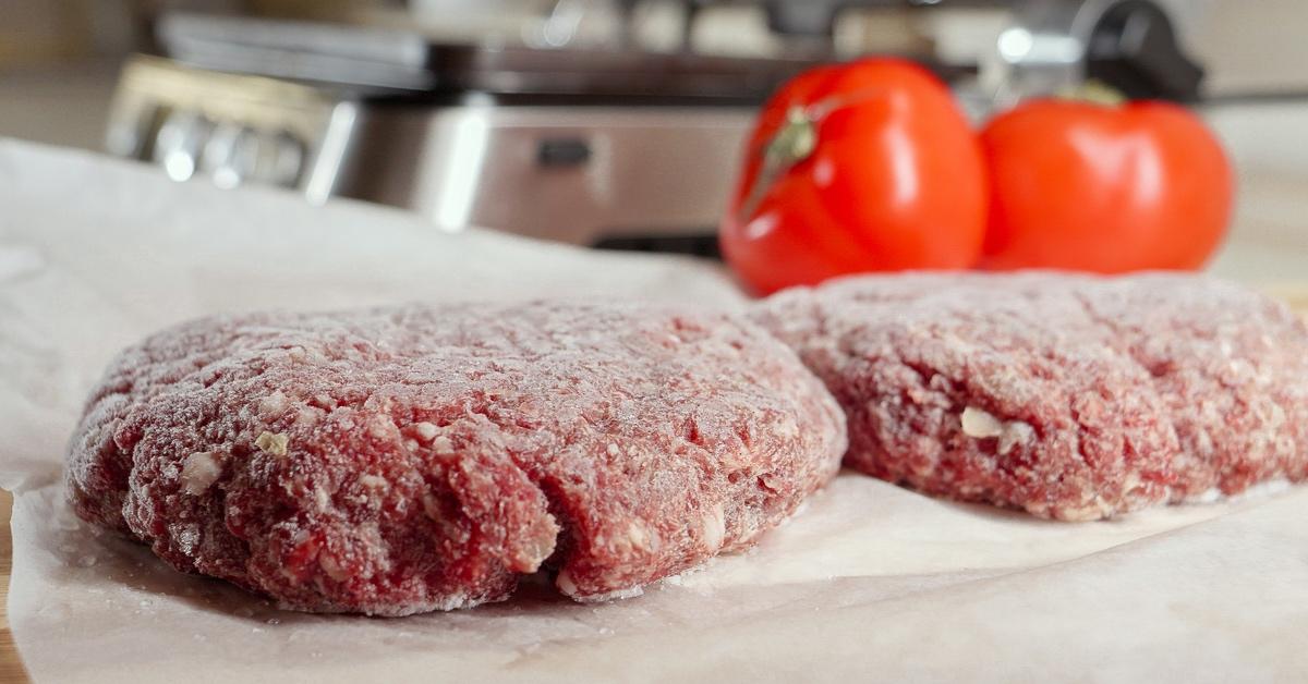 Frozen hamburger patties on a piece of parchment paper next to whole tomatoes.