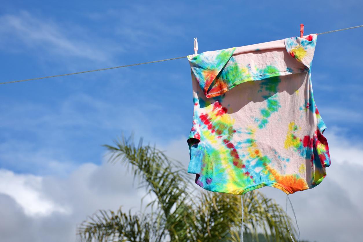 A tie-dye shirt hanging upside down on a clothesline outside with a blue sky and a green palm tree in the background.