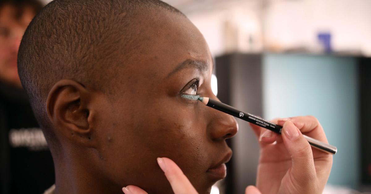 Model looks upwards as a makeup artist's hands apply green eyeliner below the model's eye