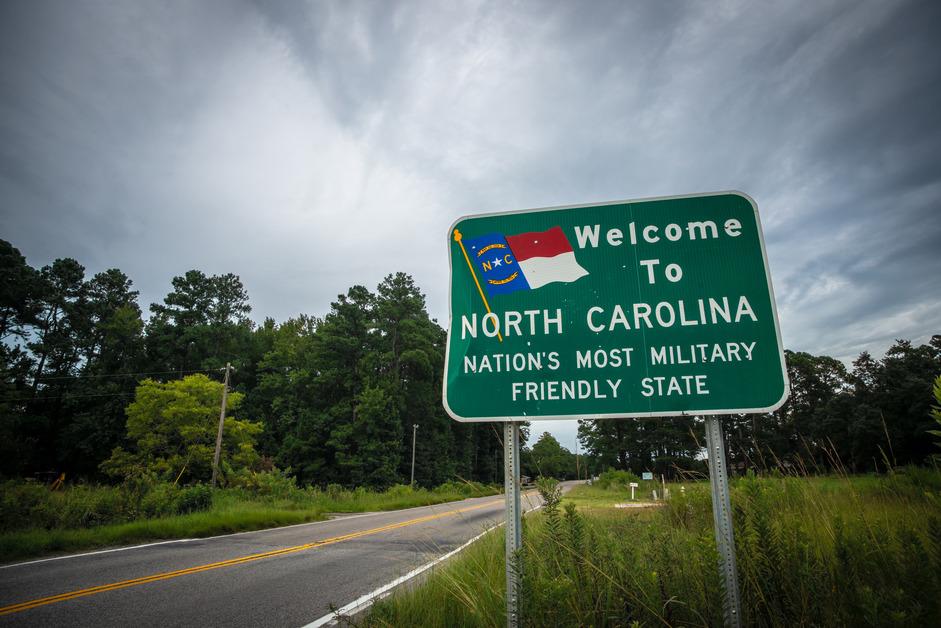 A green highway sign that says, "Welcome to North Carolina, Nation's Most Military Friendly State," with a continuing road in the background in a rural setting. 