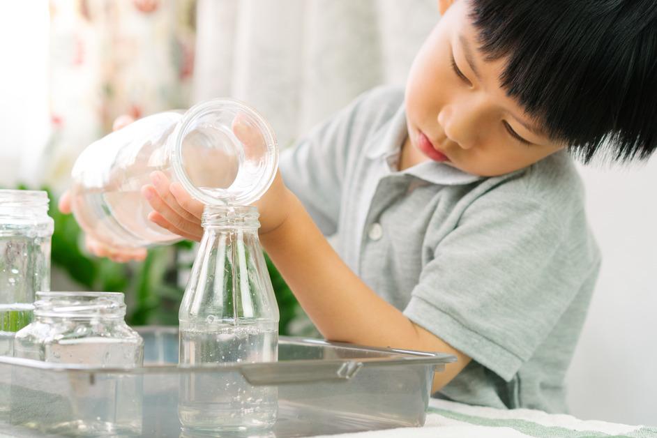 A boy focuses on pouring water from one glass container into another surrounded by other clear containers of water. 