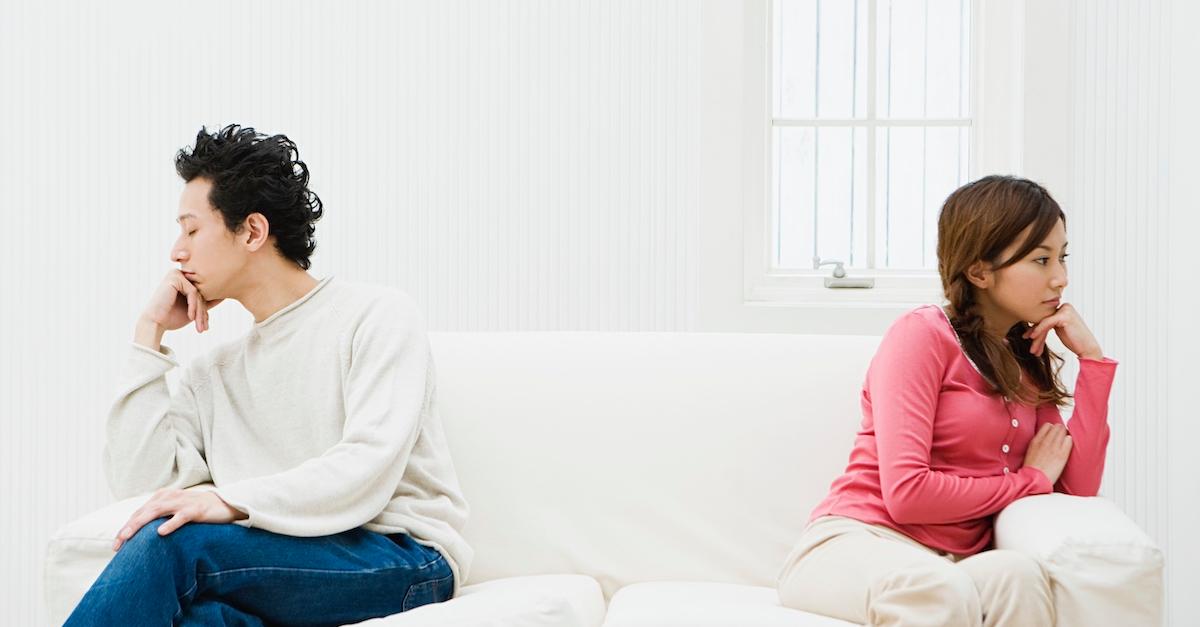 Man and woman sit on opposite sides of a white couch, looking away from each other angrily