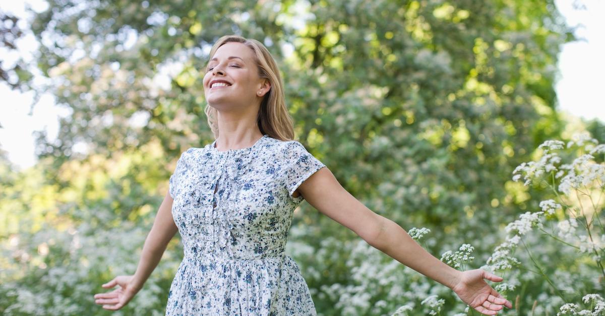 A blonde woman stands in a garden with her eyes closed and arms spread.