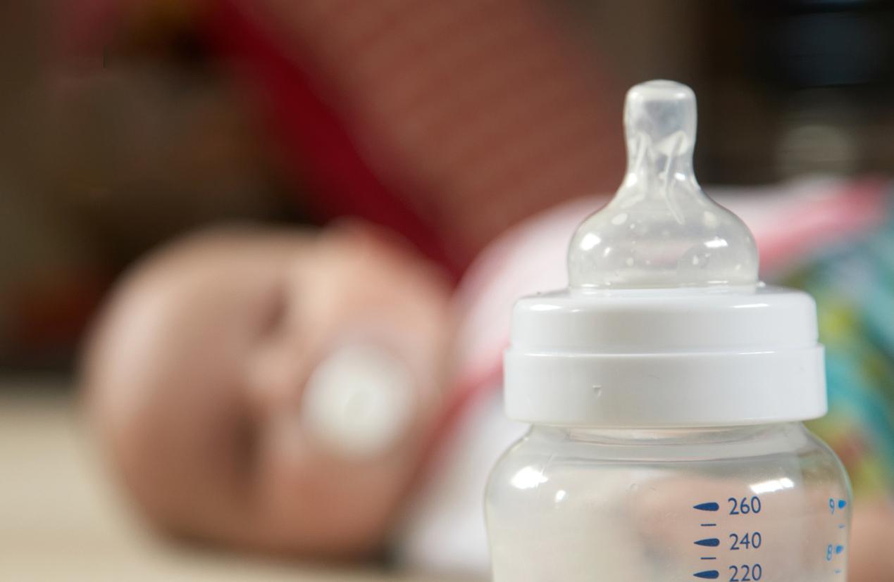 A plastic baby bottle appears in the foreground while a baby is blurred in the background.