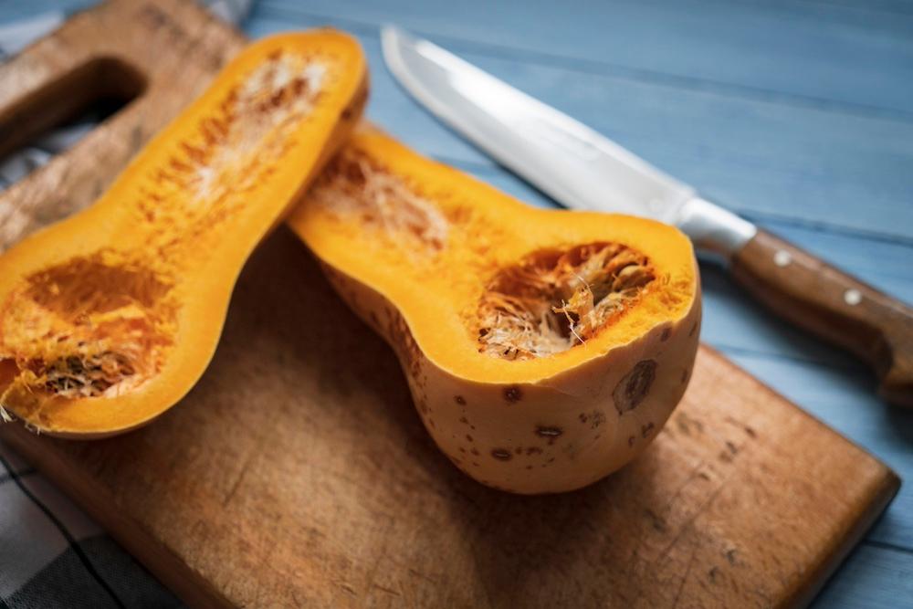 A butternut squash cut in half next to a knife and on top of a cutting board.