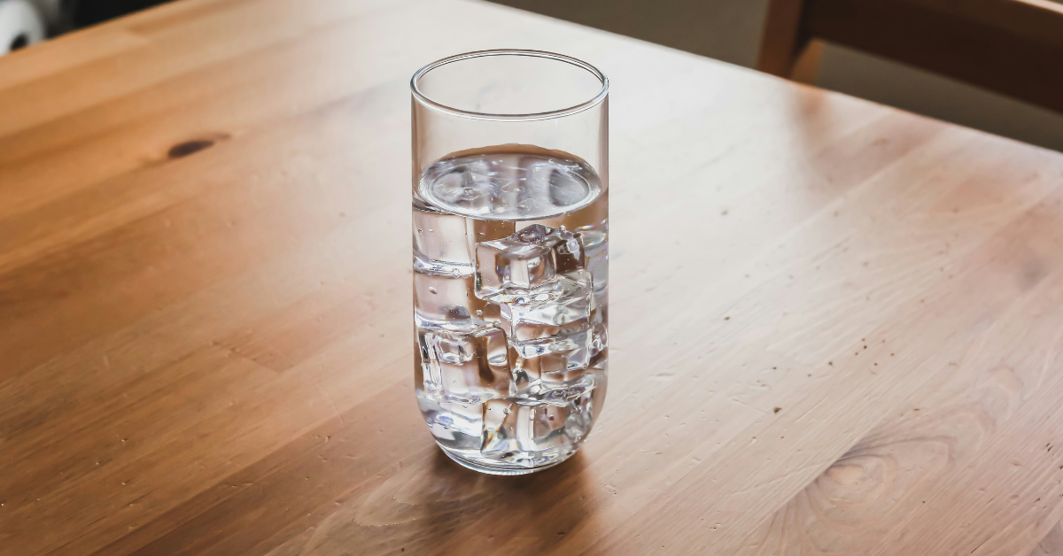 A glass of water filled with ice sits atop a wooden kitchen table