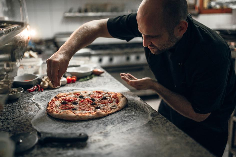 A chef standing in a kitchen adding toppings to a pizza. 