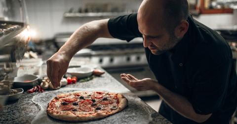 A chef standing in a kitchen adding toppings to a pizza.