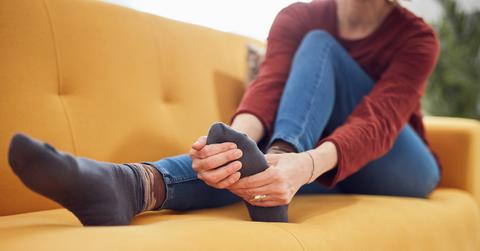A person wearing a red shirt, jeans, and dark socks sits on a yellow couch and grabs their foot.