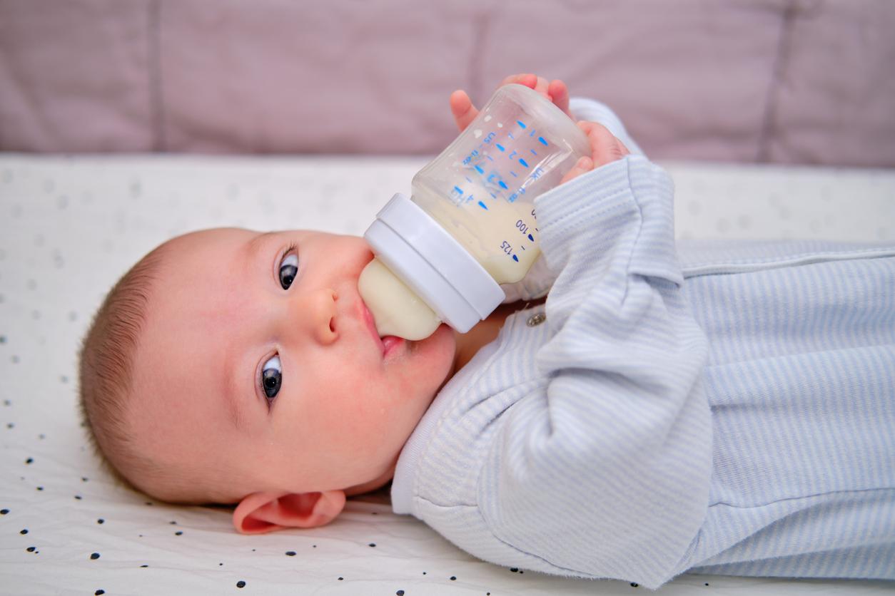 A newborn baby suckles from a plastic baby bottle while laying in a crib.