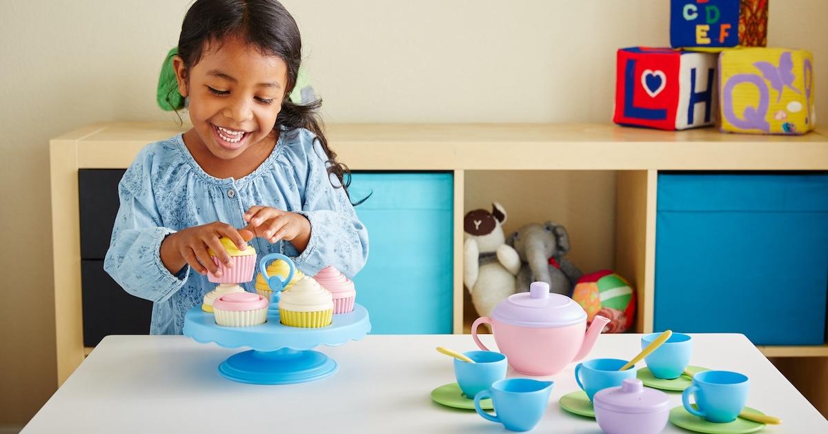 Young girl smiles at table while playing with toy cupcakes and tea set.