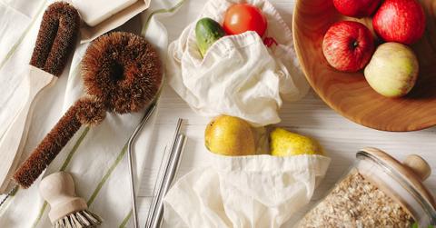 Taken from above, on a white table, are various fruits in a bowl and white cloth bags, metal straws, and other zero-waste items.