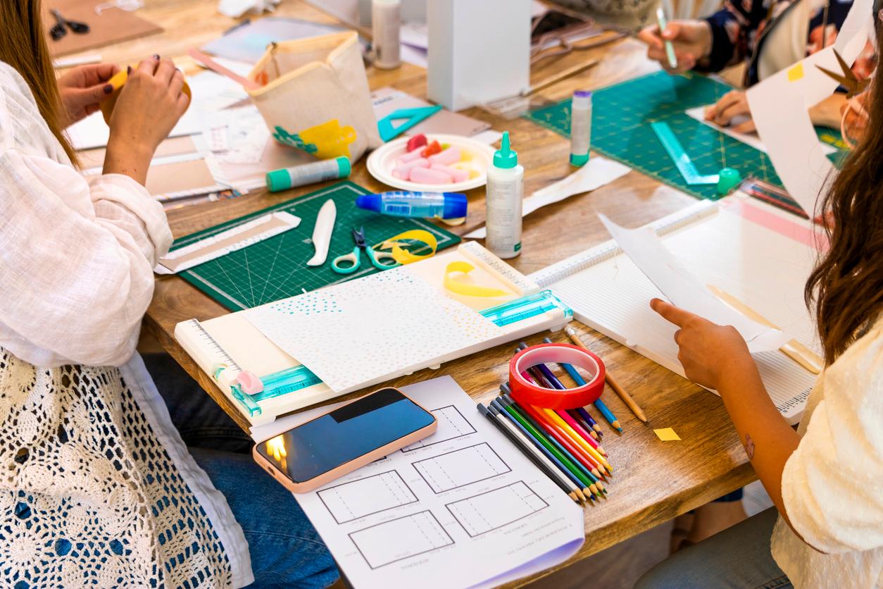 People scrapbooking at a large table covered in supplies like tape, colored pencils, paper, scissors, and glue.