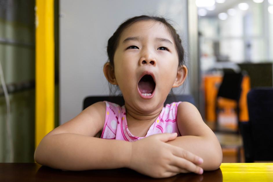 Young girl wearing a pink tank top sits at a table and yawns. 