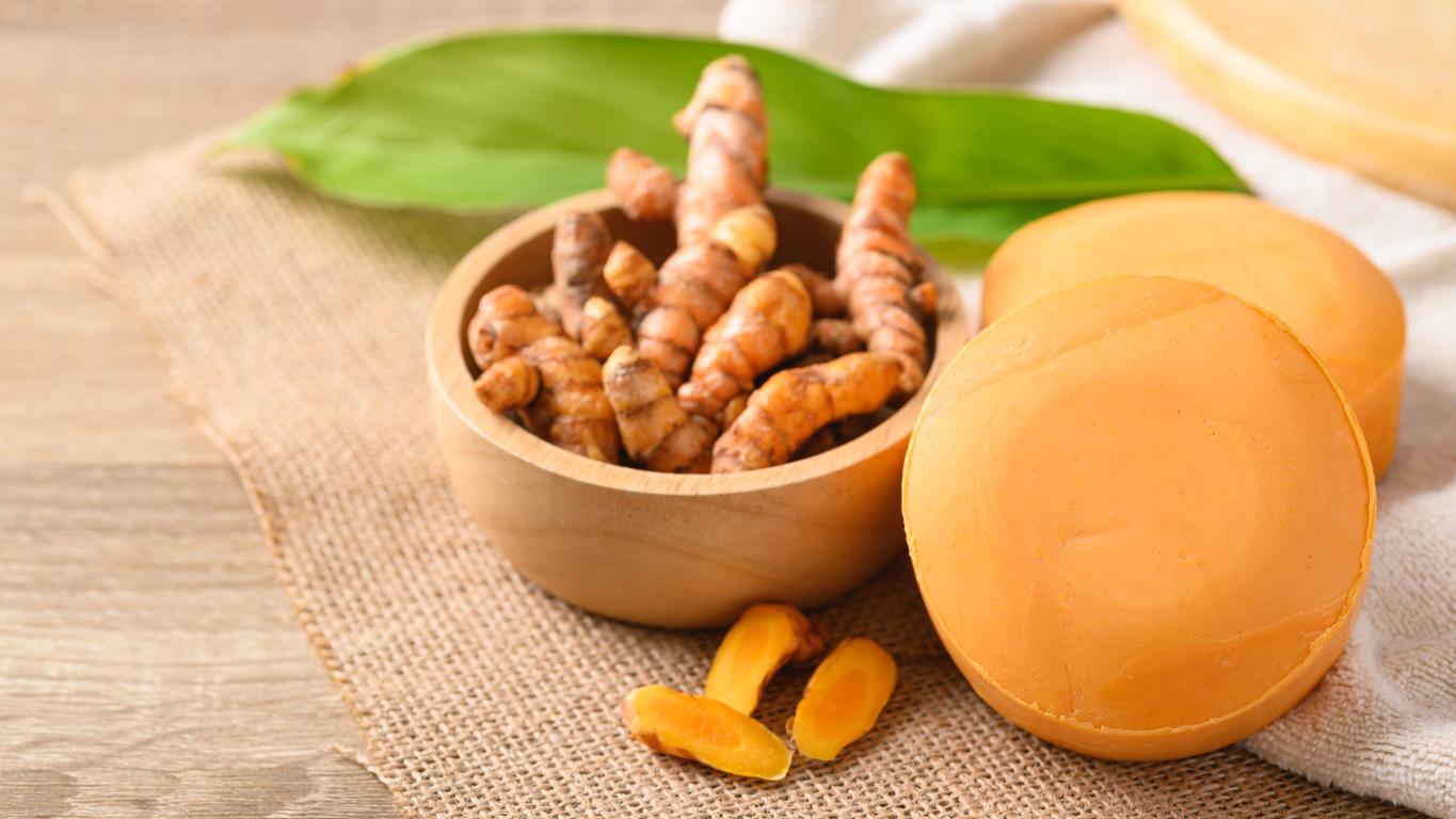 Turmeric root is pictured in a wooden bowl atop a brown cloth beside two turmeric-based soaps.