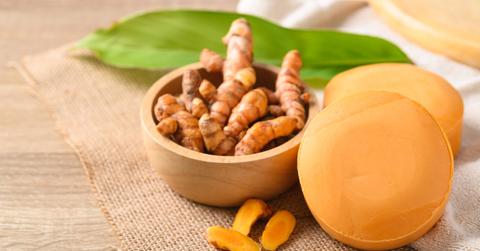 Turmeric root is pictured in a wooden bowl atop a brown cloth beside two turmeric-based soaps.