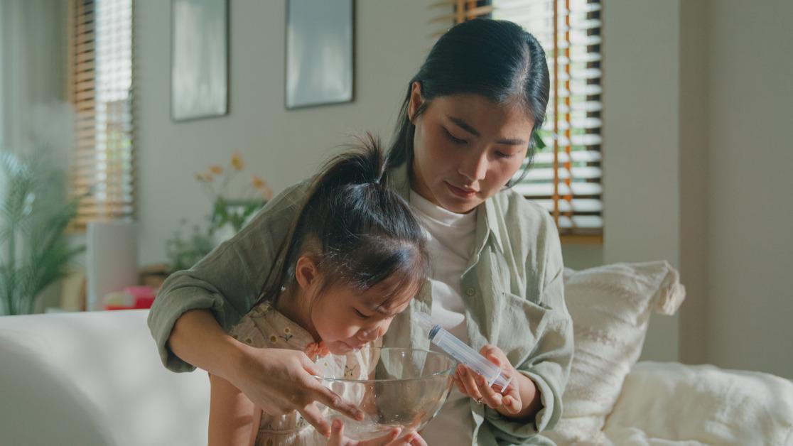 A mother and daughter sit on the couch while the mother holds a nasal syringe above a bowl near the girl's face. 