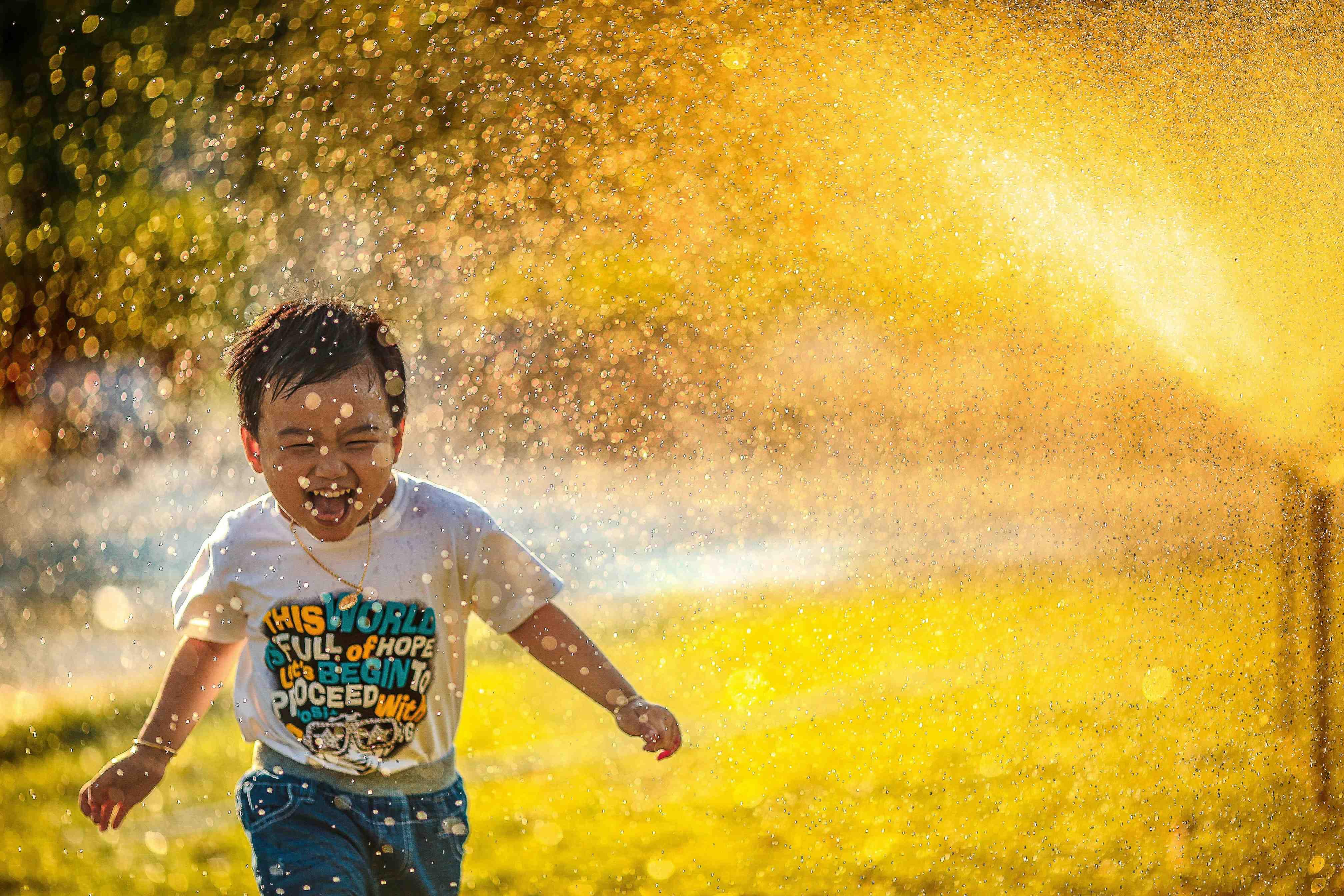 A young child smiles as he plays in the grass with a water sprinkler.