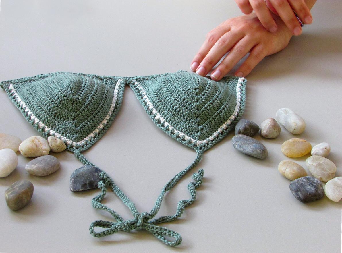 A green crocheted triangle swimsuit top on a white table next to some small rocks.