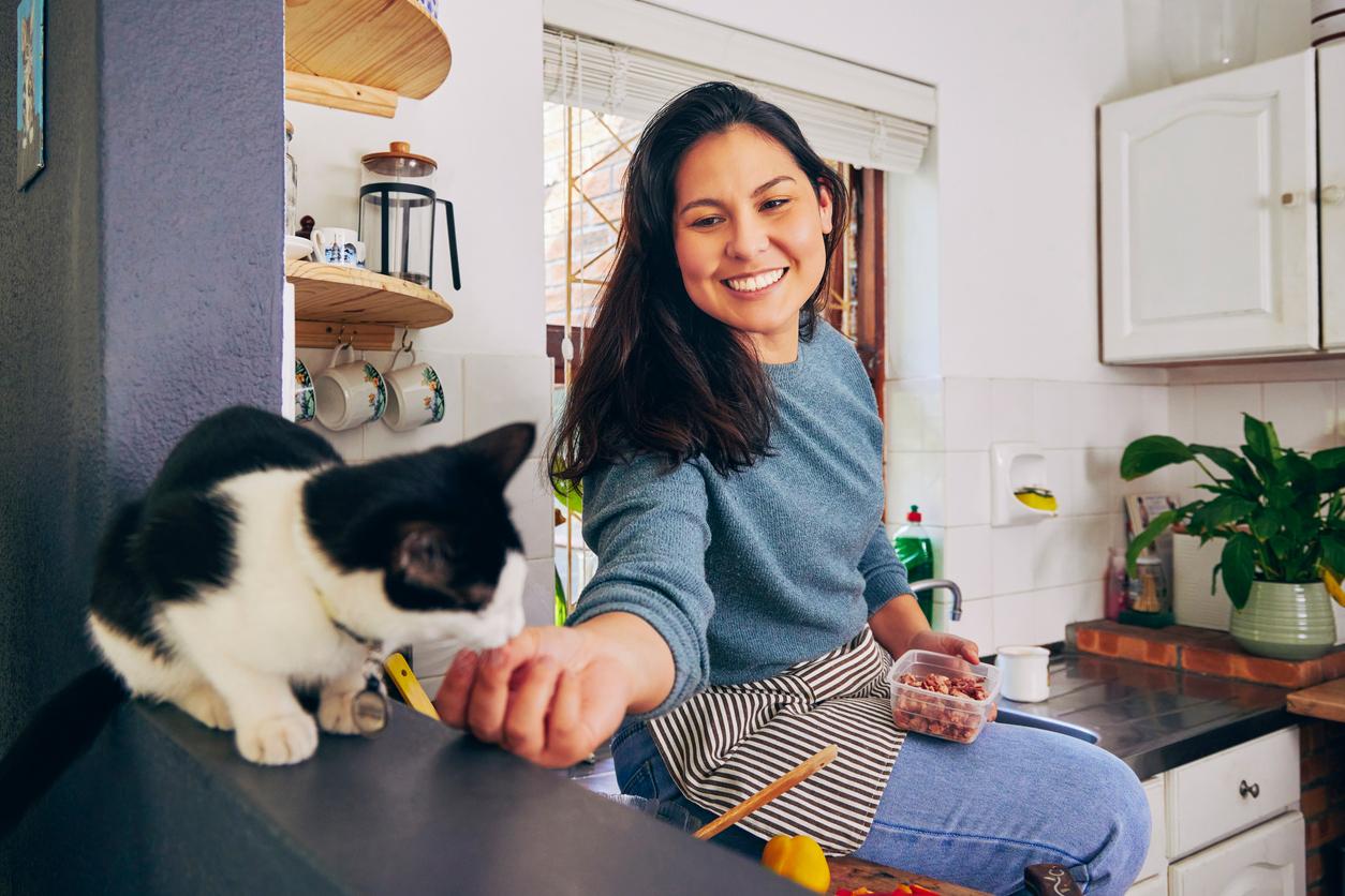 A smiling woman sits on top of a kitchen counter and feeds her black-and-white cat bacon bits.