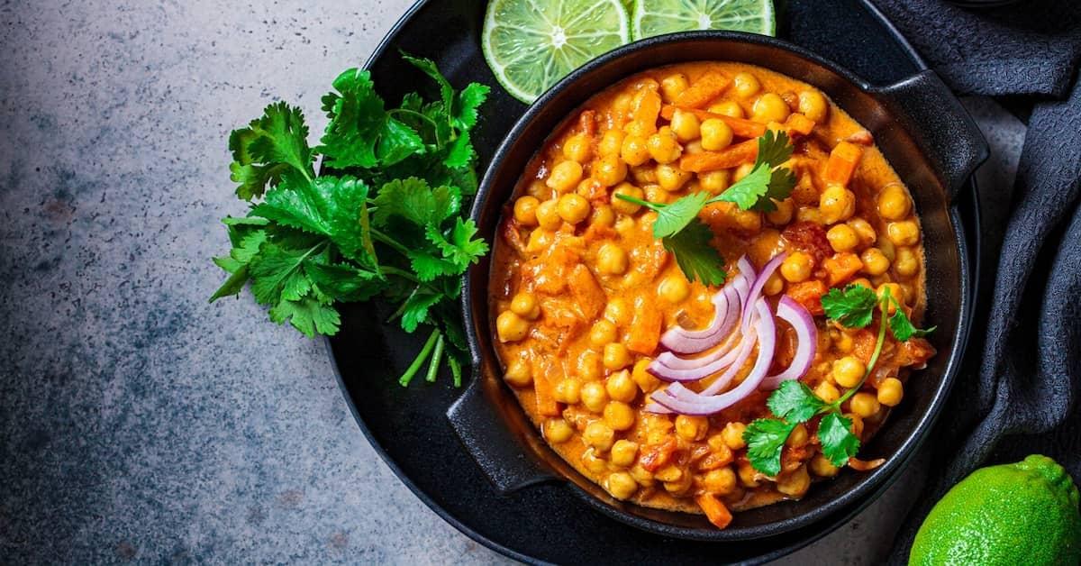 Vegan chickpea curry garnished with cilantro, red onion slices, and lime, in a black bowl