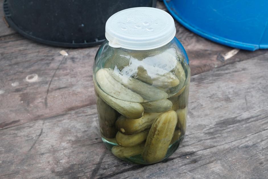 A large glass jug of pickled cucumbers with a plastic lid on a table.