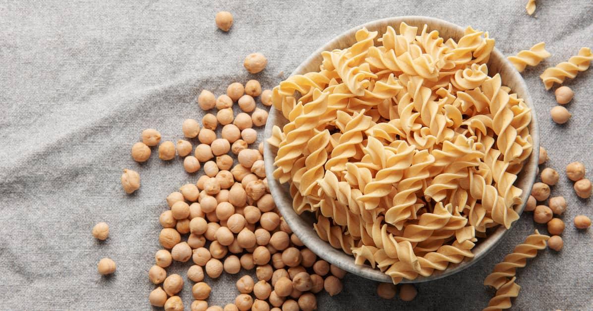 Uncooked chickpea pasta in a ceramic bowl with chickpeas scattered on a grey tablecloth.