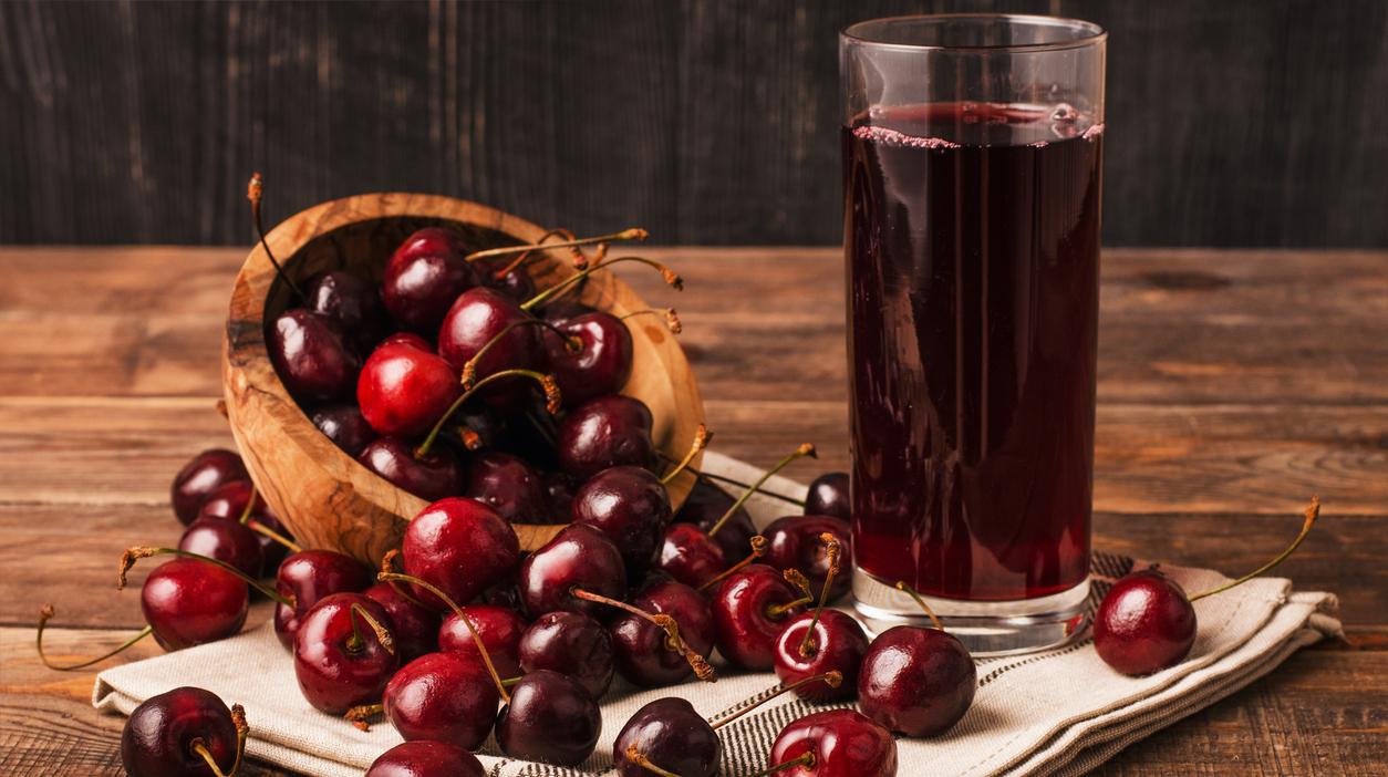 Cherry juice in a tall glass next to a wooden bowl of half spilled cherries on a table. 