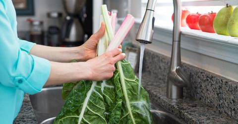 A woman wearing a blue shirt washes collard greens in a kitchen sink.