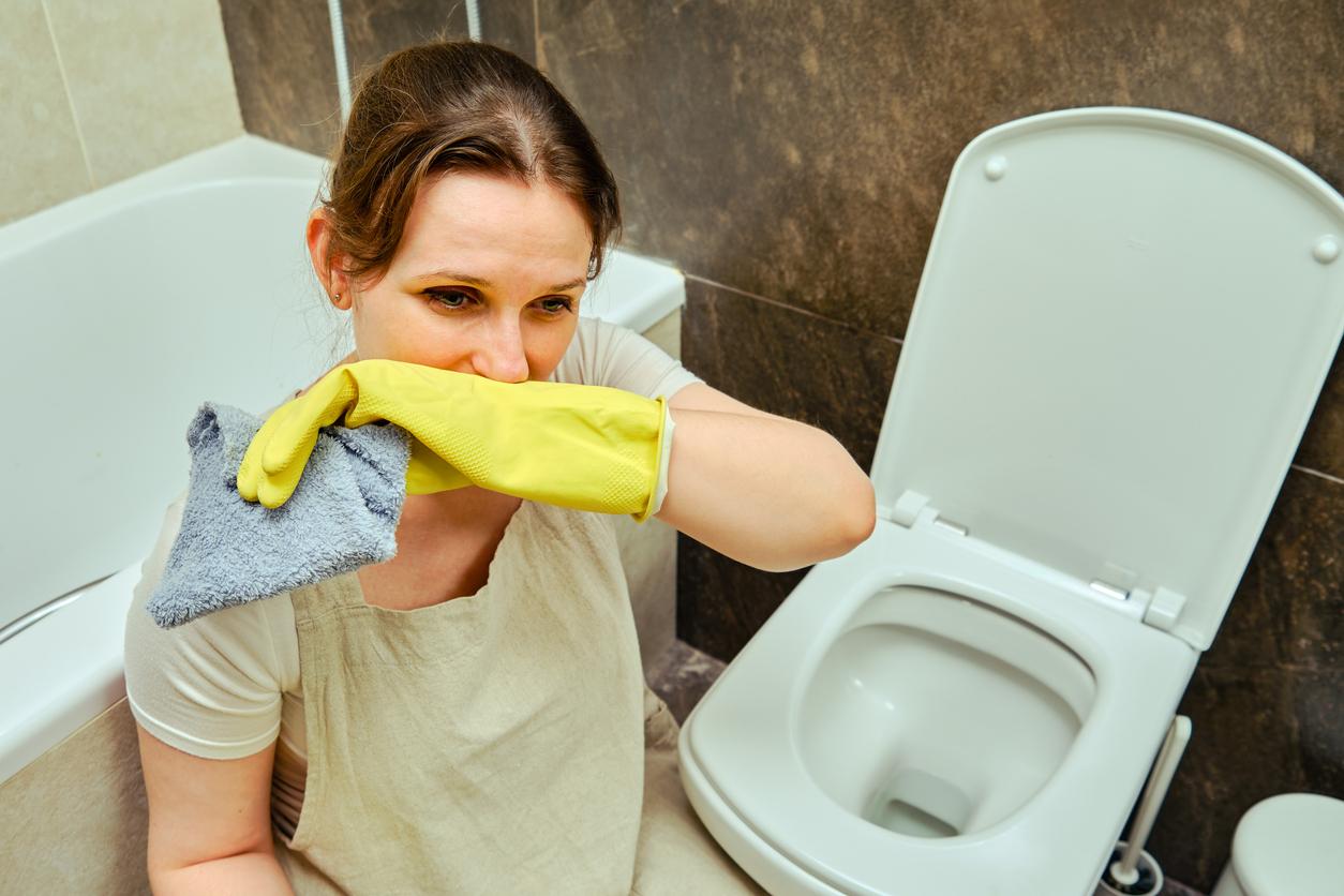 A woman wears yellow rubber gloves while holding a grey rag above a toilet and places her wrist below her nose.