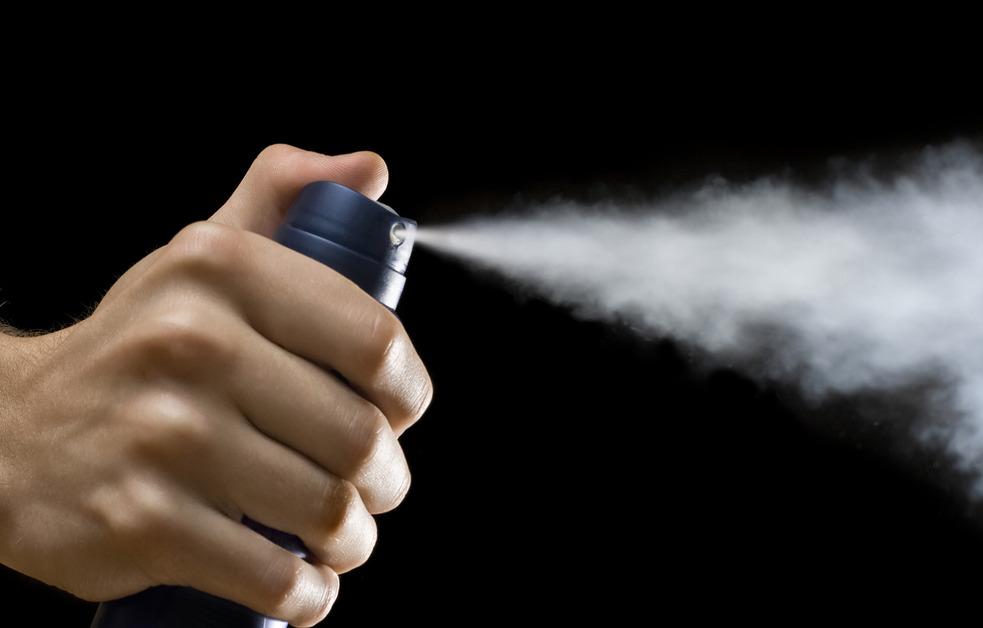 A close up of a person's hand with their thumb pressing down the top of an aerosol spray can releasing it's contents in front of a black background. 