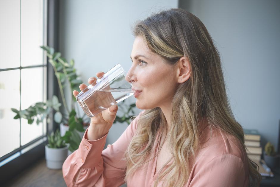 Side view of a blonde woman in pink blouse drinking a glass of water while looking out the window. 