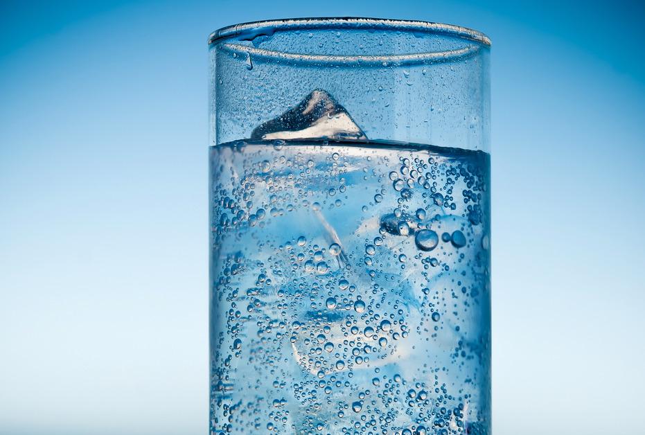 A clear glass of a bubbling drink with ice in front of a blue background. 
