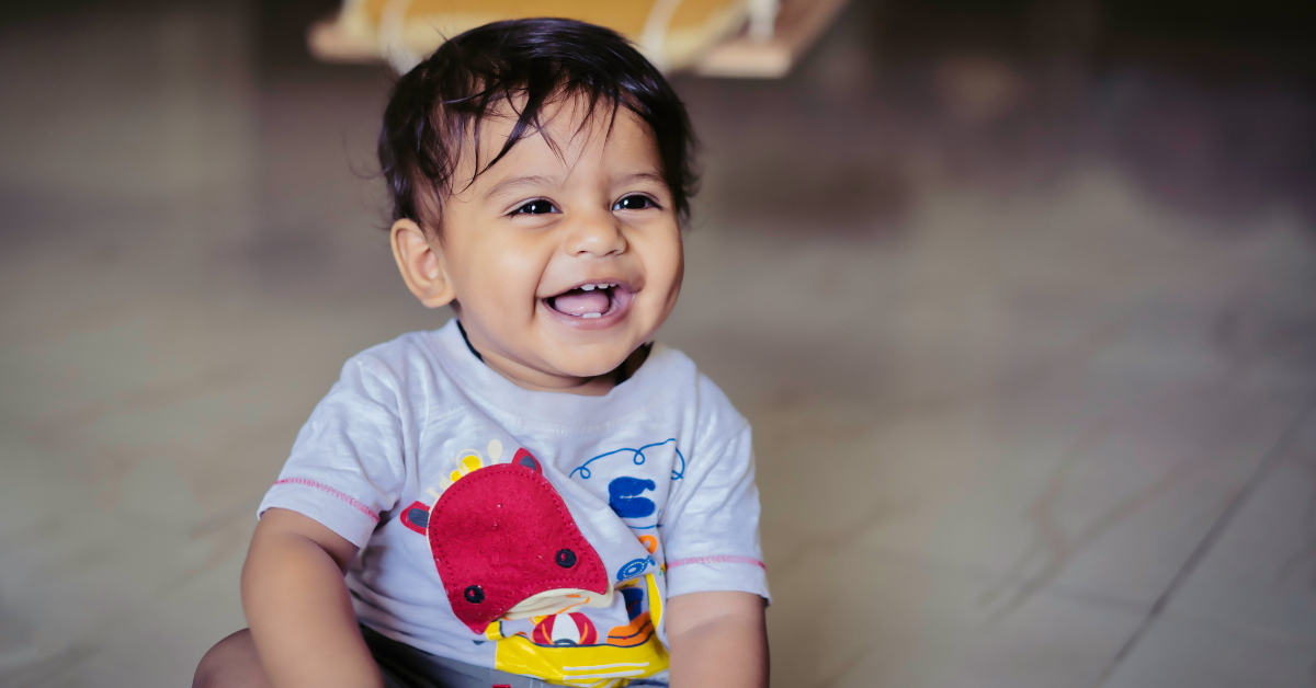 A toddler smiles with a full toothy green as he sits on the floor