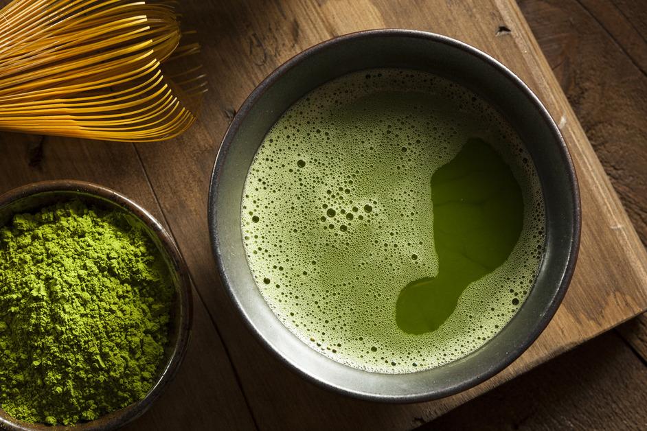 A cup of match sits on a wooden surface next to to a bowl of matcha powder. 