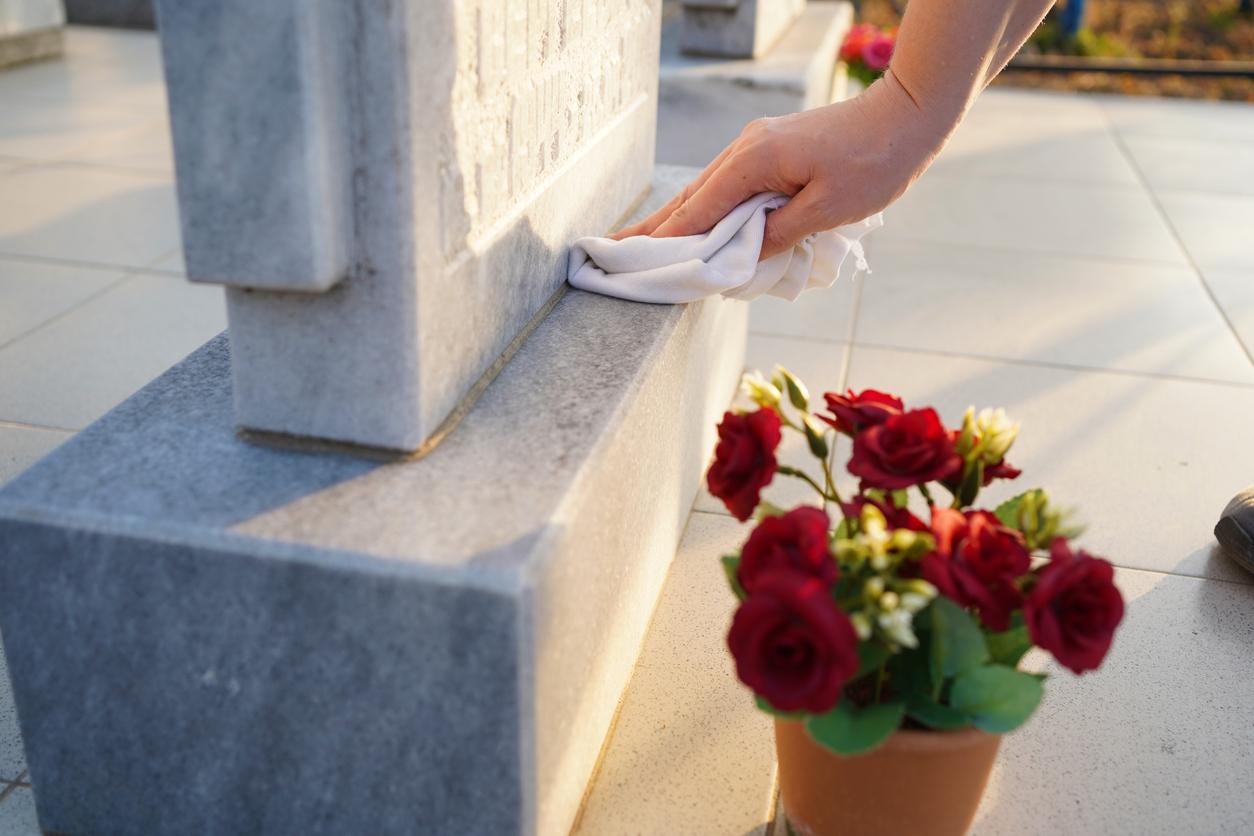 A person wipes a headstone with a white cloth beside a pot of flowers.