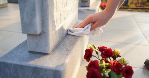 A person wipes a headstone with a white cloth beside a pot of flowers.