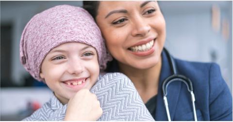 A young sick girl with a pink head covering smiles next to a female doctor.