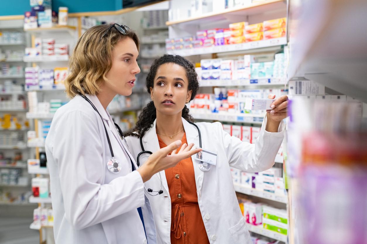 Two medical professionals wearing white coats in a pharmacy look concerned as they view medications.